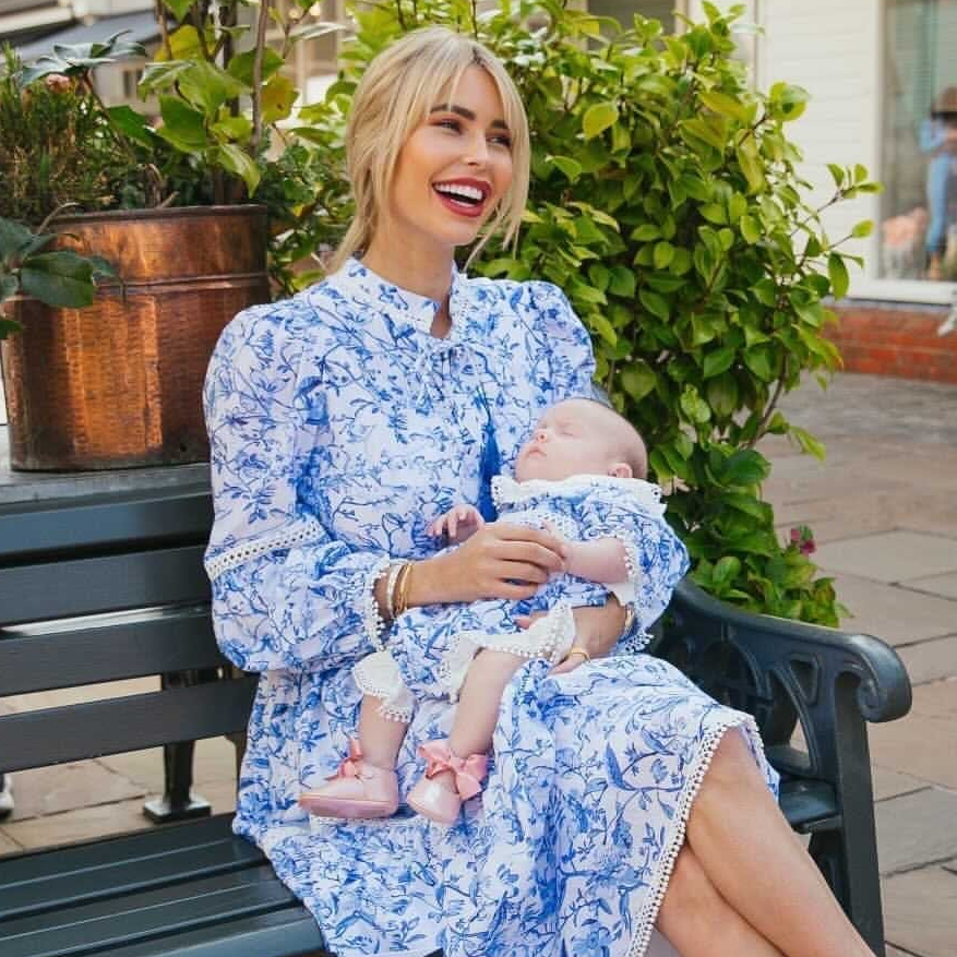 Woman in a blue floral dress holding a baby on a bench outdoors.