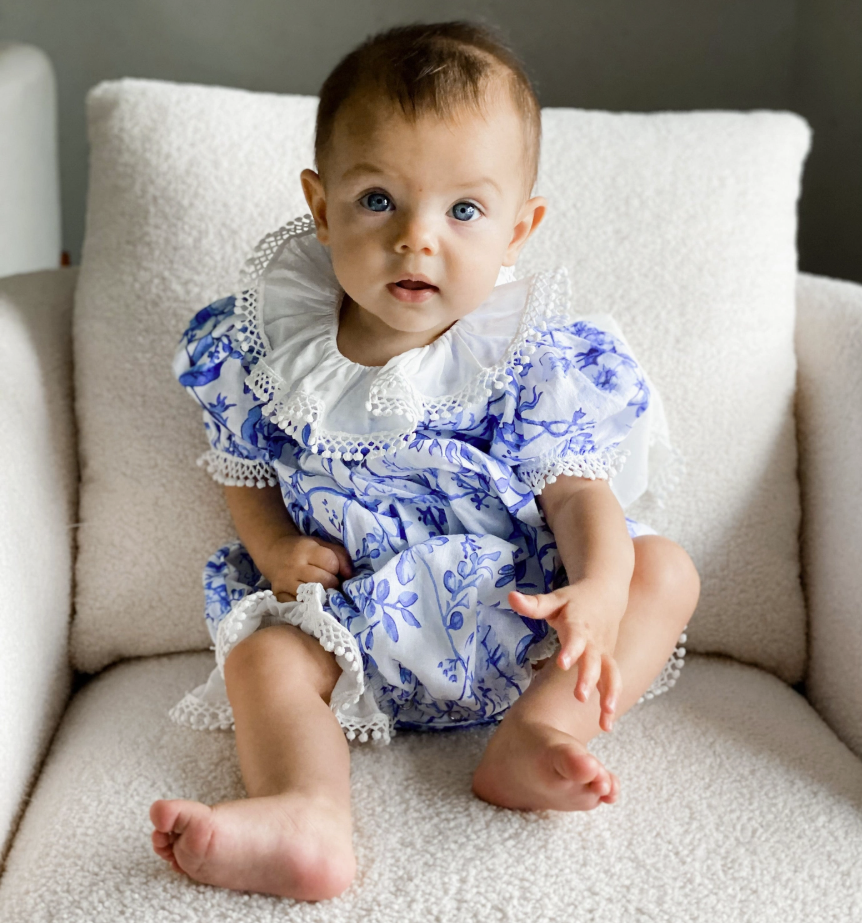 Baby in a blue and white floral dress sitting on a beige couch.