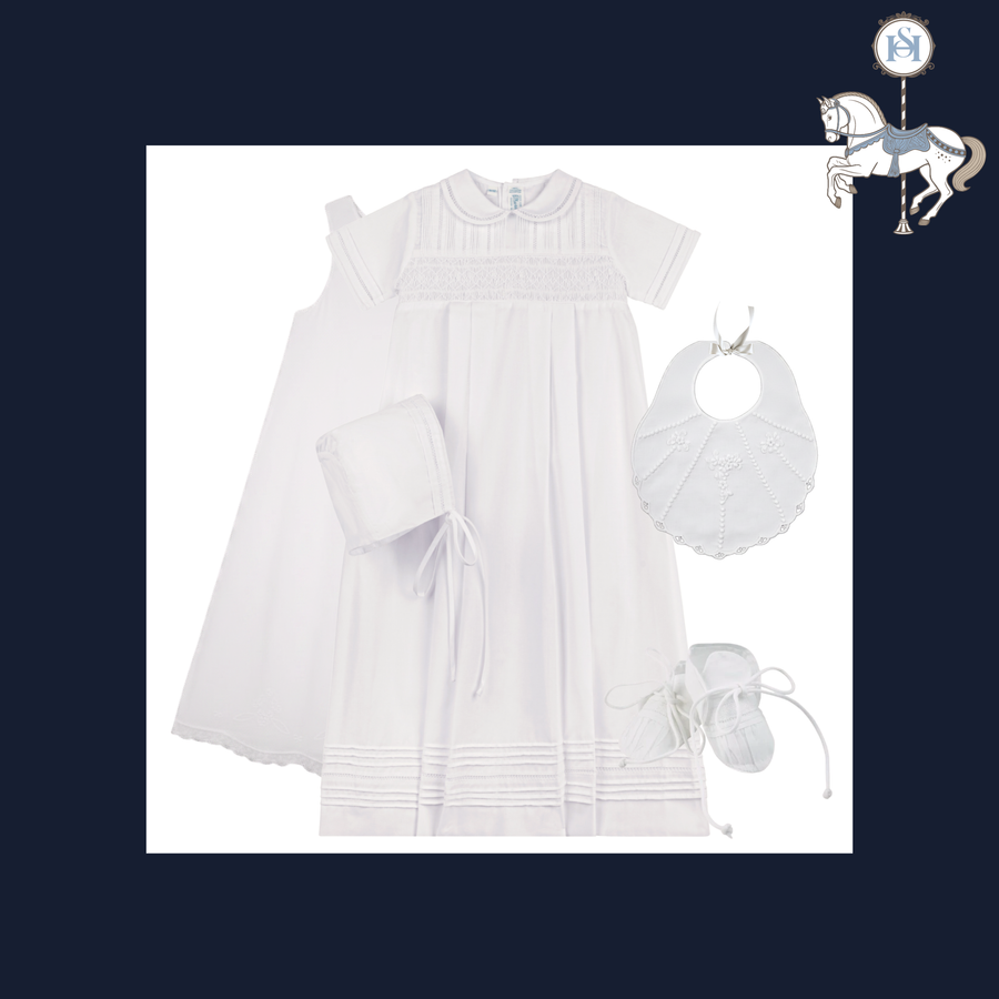 A white baby first communion outfit set featuring a short-sleeve smocked dress, a matching bonnet, a bib, and baby booties, arranged on a white background with a navy blue border and a small carousel horse logo in the top right corner.